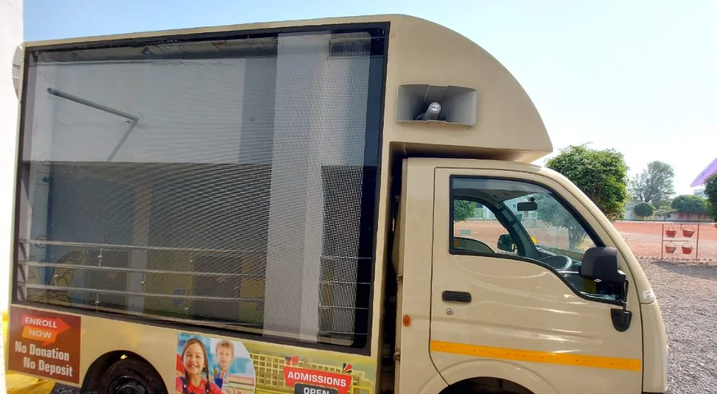 A cream LED advertising van with a large side-mounted digital display and roof loudspeaker, parked outdoors in daylight